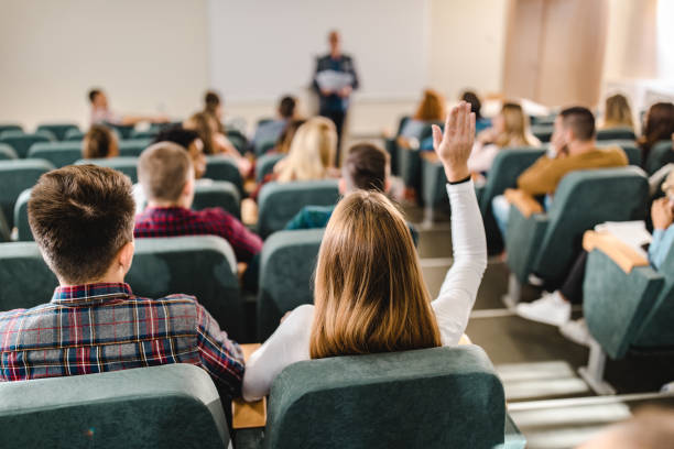 Rear view of a female college student raising hand in amphitheater. Back view of female college student raising her hand to answer the question on a class at lecture hall. hand raised classroom student high school student stock pictures, royalty-free photos & images