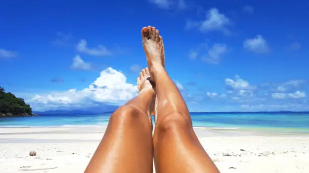 Woman tanned skin legs sunbathing on the white sand beach with the blue sky and sea in the background, Summer holidays and vacations concept.