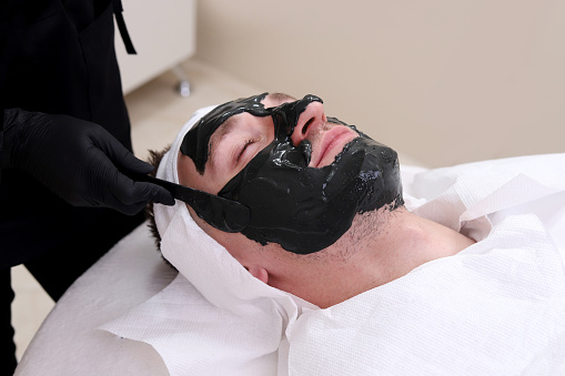Cleaning the face of a man in a beauty salon. Spa therapy for men receiving facial black mask.