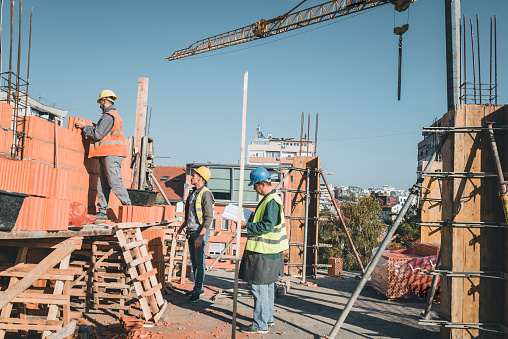 Teamwork by Three Builders with Protective Helmets who are Building a Wall of Bricks at the Building Site. Young Building Workers is Working Together at the Roof of the Construction Site Under Control of Their Experienced Team Leader.