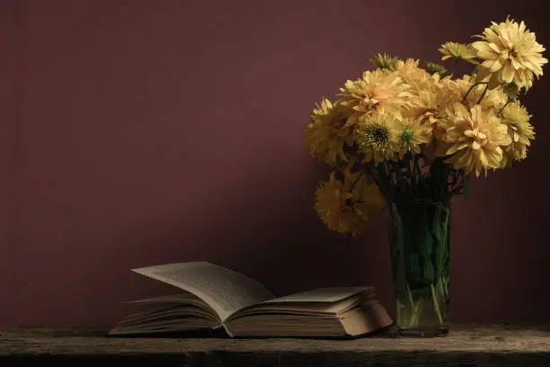 Old book and yellow flowers in vase on a oak wooden table, dark-red wall background.