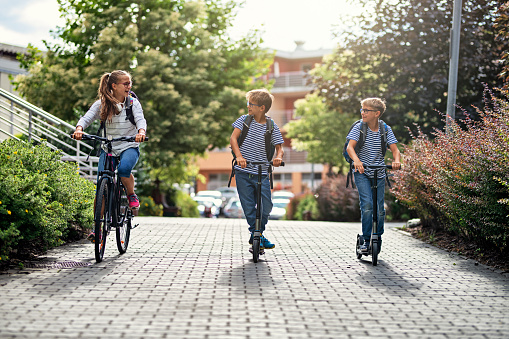 Three kids riding to school. The teenage girl is riding a bike and two brothers are riding scooters.\nNikon D850