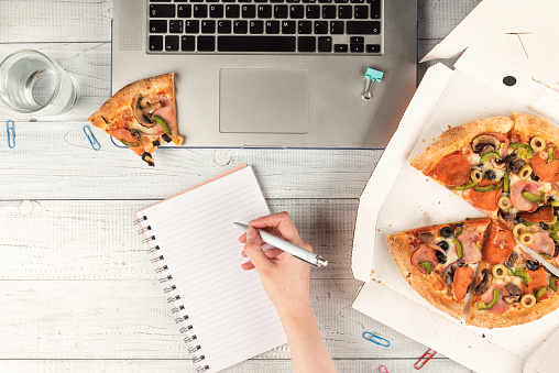 Businesswoman working an office desk with a laptop and having a lunch break with a tasty pizza and glass of water. Top view, flat lay