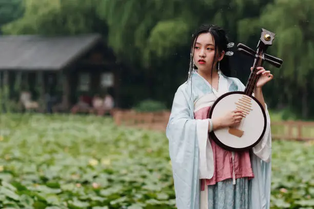 Beautiful Asian woman in cyan costume clothes hanfu holding yueqin (four stringed plucked instrument) with lotus pond background in rainy day, traditional ancient Chinese beauty, time travel fiction.