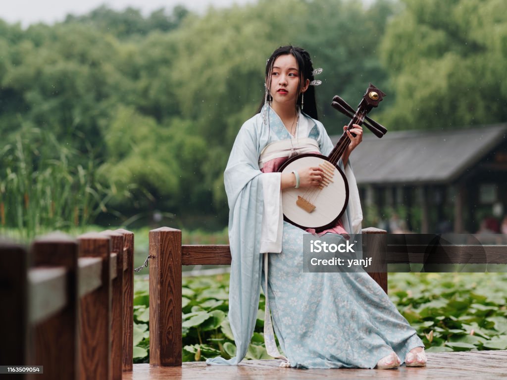 Beautiful Asian woman in cyan costume clothes hanfu holding yueqin (four stringed plucked instrument) with lotus pond background in rainy day, traditional ancient Chinese beauty, time travel fiction. Hanfu Stock Photo