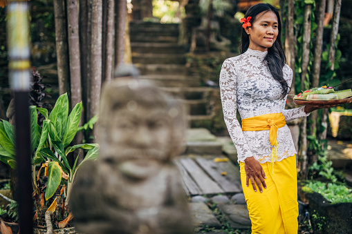 One woman, young lady in traditional Balinese clothing standing outdoors, holding a plate with flowers.