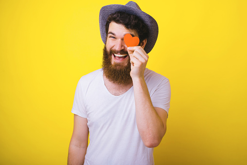 Portrait of romantic guy with beard and hat, covering eye with little paper heart, standing over yellow background