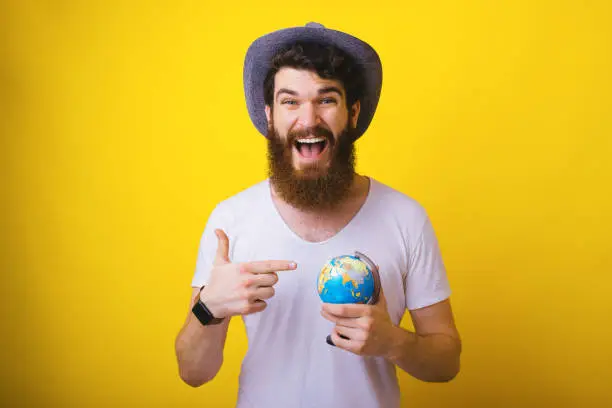 Photo of bearded man, waring hat, excit for new trip, pointing at little globus, over yellow background