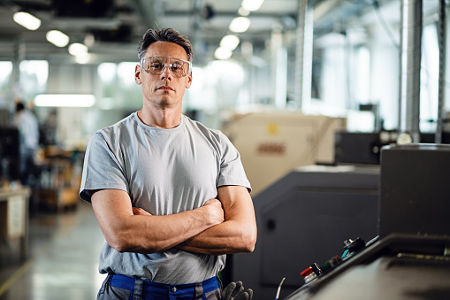Portrait of industrial engineer with arms crossed standing by CNC machine and looking at camera.
