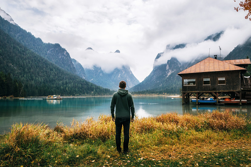 Young man hiker stands back to camera on shore of Dobbiaco Lake or Toblacher in Dolomites with wooden boathouse hut, Italy