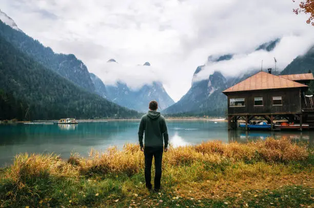 Photo of Young man stands back to camera on shore of Dobbiaco Lake or Toblacher in Italy