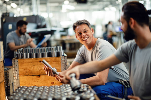 Happy manual workers talking while working together at steel factory. Two happy steel workers preparing manufacture products for the distribution and communicating in industrial facility. Focus is on mid adult worker. manufacturing occupation stock pictures, royalty-free photos & images