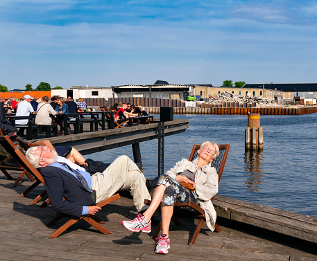 Two people sound asleep in deck chairs beside the harbour in Copenhagen, Denmark’s capital. Other people enjoy drinks or socialising on the jetty in front of the Royal Danish Playhouse (Skuespilhuset) in the Frederiksstaden district of the city.