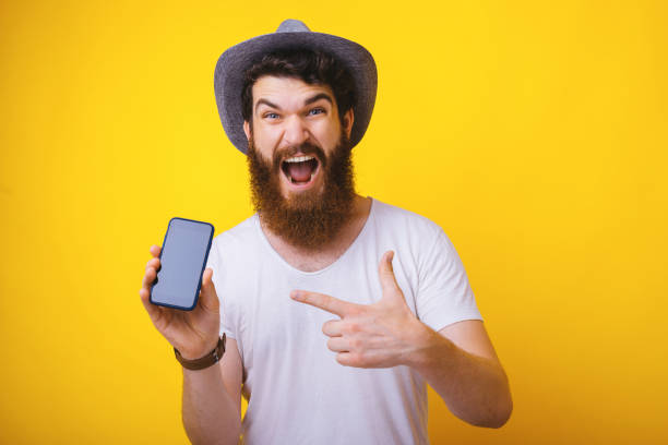istock Photo of excited bearded guy, wearing hat, screaming at camera while pointing at mobile screen, standing over yellow background 1163522117