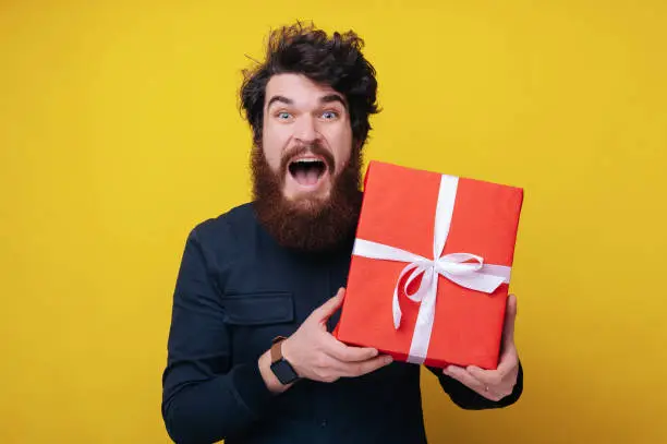 Photo of Handsome bearded man, looking excited at camera, holding a gift box, and standing over yellow background