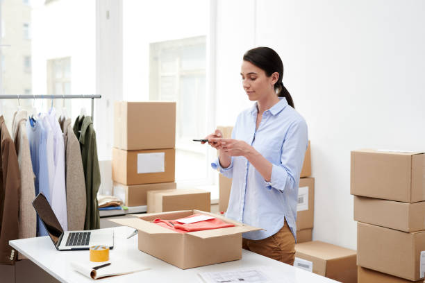 istock Young female manager with mobile phone photographing packed clothes 1163522794