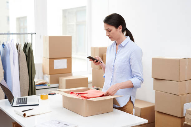 istock Young mobile female with smartphone taking photo of folded pullover in open box 1163522796