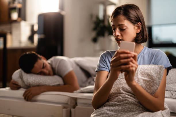 istock Young woman text messaging on mobile phone while her boyfriend is sleeping on the bed. 1163533799