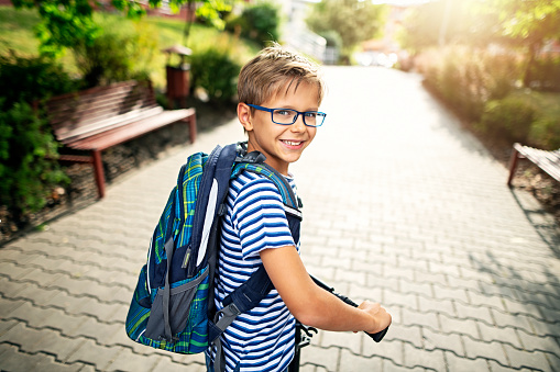 Little boy wearing backpack is riding to school on his push scooter.\nNikon D850