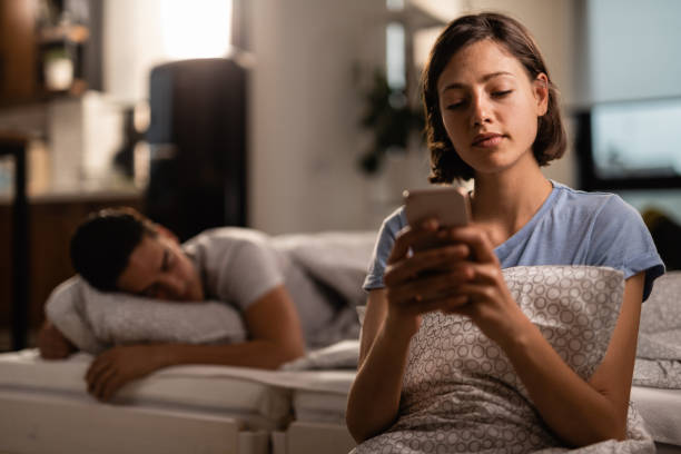 Young woman using smart phone in bedroom while her boyfriend is sleeping. Young woman text messaging on cell phone while her boyfriend is sleeping on the bed behind her. infidelity photos stock pictures, royalty-free photos & images