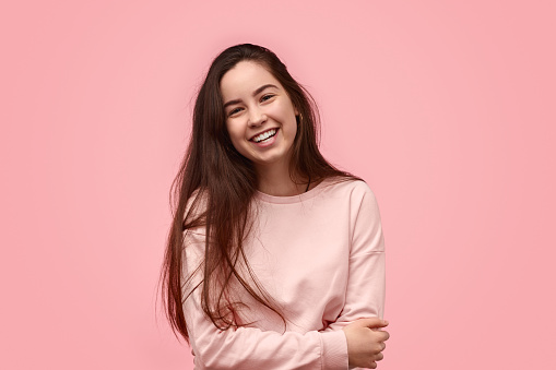 Happy teen girl with long hair laughing and embracing torso while standing against pink background