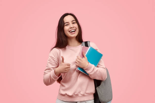 Cheerful student gesturing thumb up Happy teen girl with backpack and notebooks smiling and showing thumb up gesture while approving education against pink background pink promise stock pictures, royalty-free photos & images