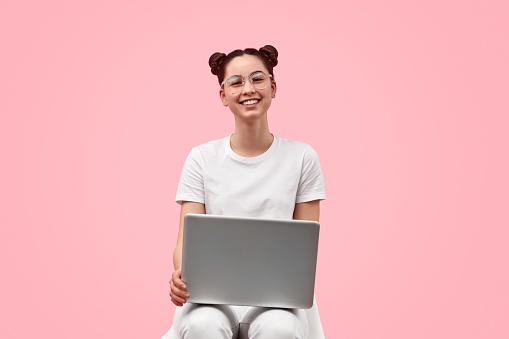 Cute teen girl in trendy glasses smiling and looking at camera while browsing modern laptop against pink background