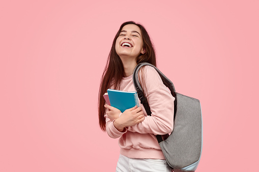 Cheerful teen girl with backpack embracing notepads and laughing while standing against pink background during studies