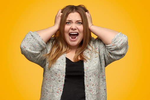 Mad plus size female in casual outfit shouting and tearing out hair while standing on yellow background and looking at camera