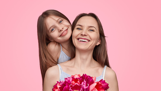 Attractive adult female with bunch of bright tulips and sweet girl cheerfully smiling and looking at camera while standing on pink background