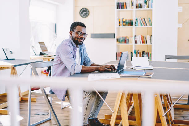 istock Cheerful african american hipster guy looking at camera while leaning in college campus using netbook, portrait of happy skilled dark skinned freelancer typing on laptop computer during working process 1163539207
