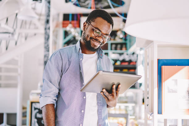istock Positive dark skinned young man in eyewear smiling getting notification about new feeds in social networks, african american blogger satisfied with online job using digital tablet in coworking space 1163539263