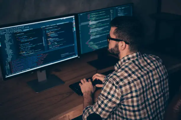 Close up back rear behind photo handsome he him his guy typing writing, keyboard development outsource IT processing language two monitors table office wear specs formalwear plaid shirt