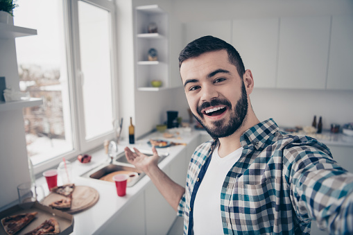 Self-portrait of his he nice attractive cheerful cheery glad bearded, guy wearing checked shirt having fun inviting you visit modern light white interior style kitchen indoors