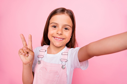 Closeup photo of little pretty lady make take selfies v-sign symbol, say hi hello friends wear overall isolated pink background