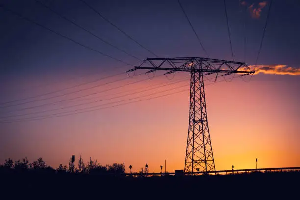 Electricity pylon with high voltage power lines silhouetted against a colorful orange and purple sunset with copy space at twilight