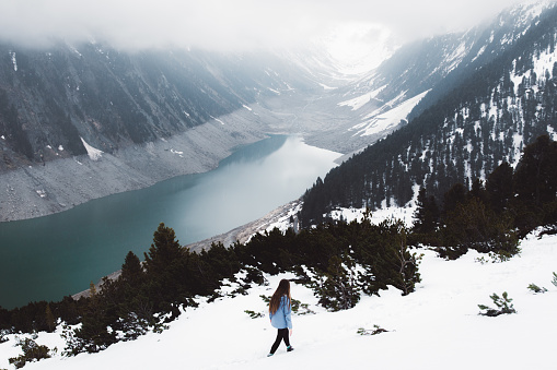 Young woman with long hair in blue jacket trekking on snowcapped Austrian Alps mountains and enjoying the view of turquoise mountain lake