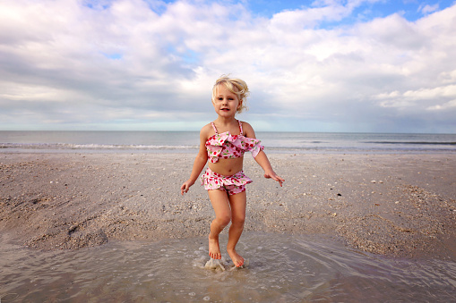 A cute little kid is jumping and playing in the water on the beach by the ocean on a summer day.
