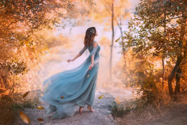 Photo of delightful light girl in sky blue turquoise dress with long flying train, princess of wind and daughter of storm, lady with dark hair throws fallen leaves to ground, autumn story in art processing