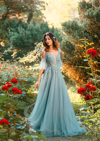 slender lady in light turquoise summer dress with cut on chest and open shoulders, princess with dark hair looks out prince in park, girl with bright smile, positive emotions, art portrait of model.