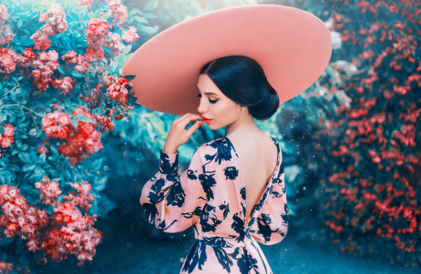 広いつばとピンクの帽子をかぶった長い黒髪から完璧な優しい髪型を持つかわいい女性、デートや写真撮影のためのエレガントな外観、オープンバックと明るい唇を持つ女性、咲くバラの女� - floral dress ストックフォトと画像
