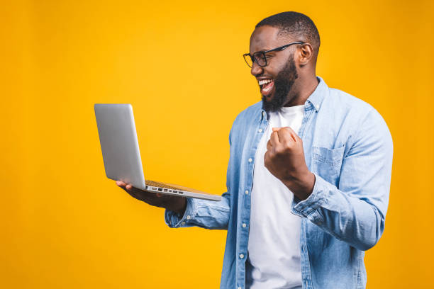 istock Excited happy afro american man looking at laptop computer screen and celebrating the win isolated over yellow background. 1163576185