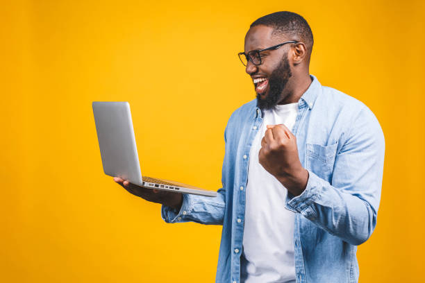 Excited happy afro american man looking at laptop computer screen and celebrating the win isolated over yellow background. Excited happy afro american man looking at laptop computer screen and celebrating the win isolated over yellow background. excitement laptop stock pictures, royalty-free photos & images