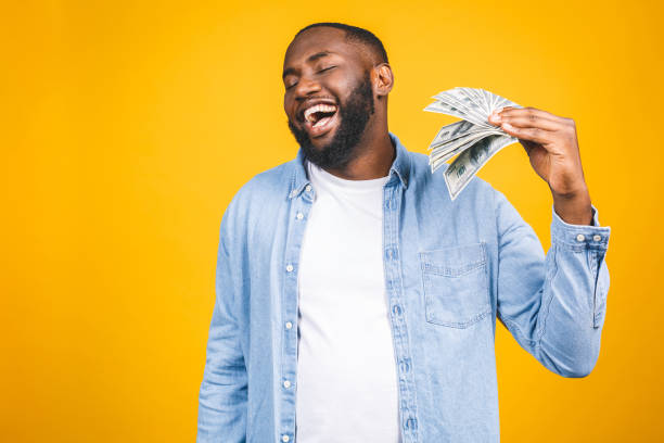 vencedor! homem rico novo do americano africano no t-shirt ocasional que prende contas de dólar do dinheiro com a surpresa isolada sobre a parede amarela. - cheerful savings men one person - fotografias e filmes do acervo