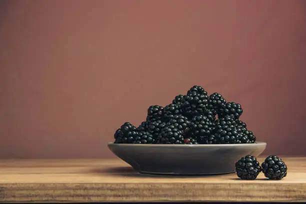 Fresh blackberry in bowl on a brown wooden table and dark-red wall background.
