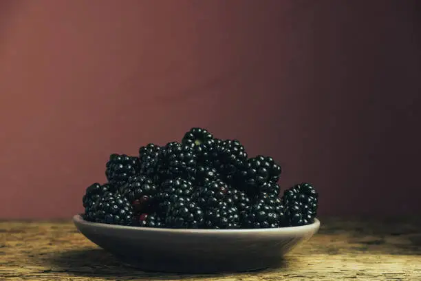Fresh blackberry in bowl on a old oak wooden table and dark-red wall background.