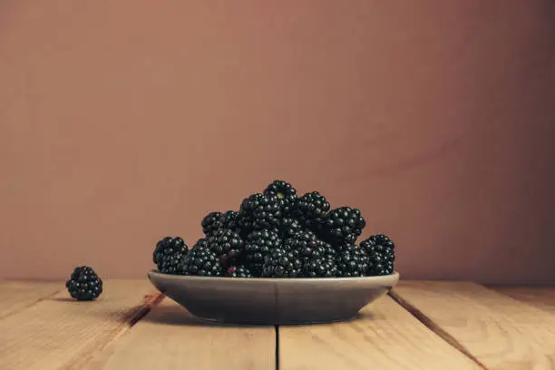 Fresh blackberry in bowl on a brown wooden table and dark-red wall background.