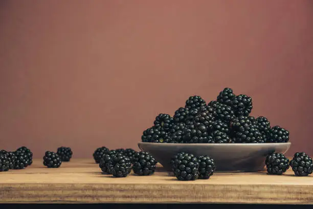 Fresh blackberry in bowl on a brown wooden table and dark-red wall background.