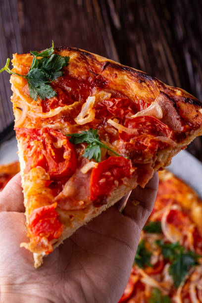istock Freshly cooked hot pizza slice holding in human hand with cheese ham meat tomato onion parsley homemade recipe on dark wooden table background close up macro 1163595663