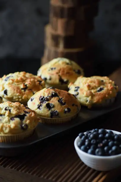 Homemade crumble top blueberry muffins with raw berries on black background. Low key still life with natural lighting side view
